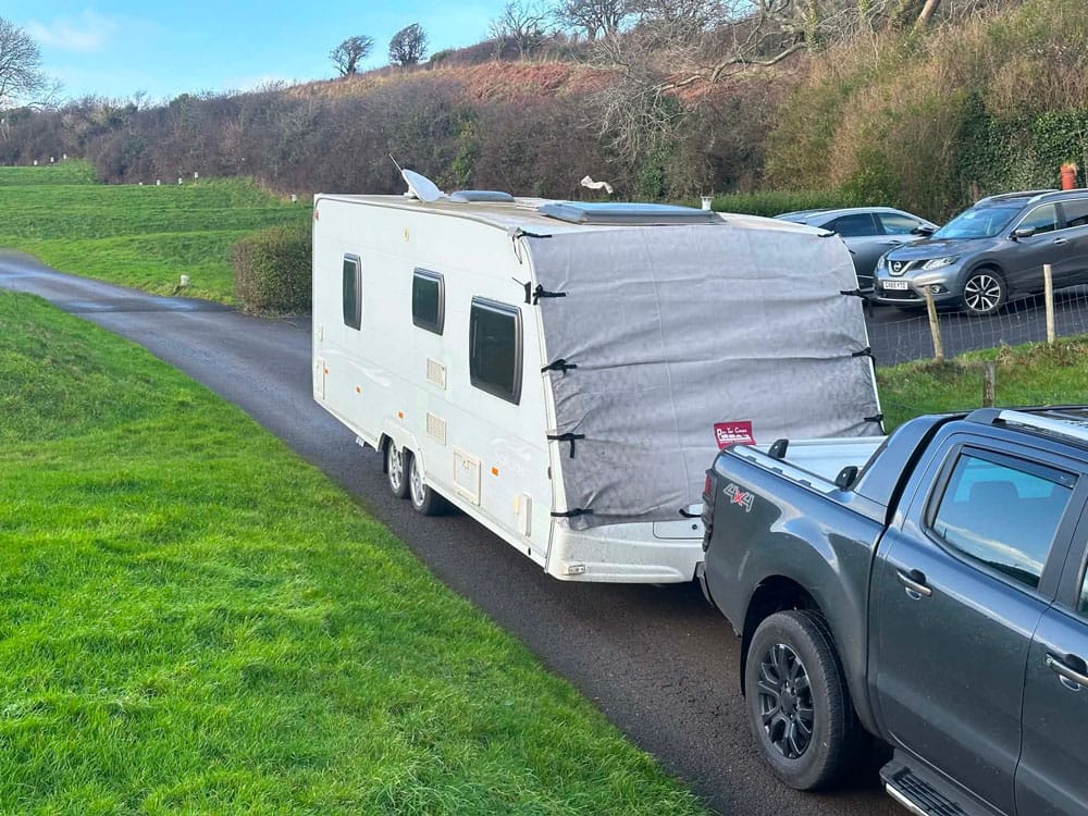 Twin-axle caravan with towing cover being collected from a country lane by a pickup truck